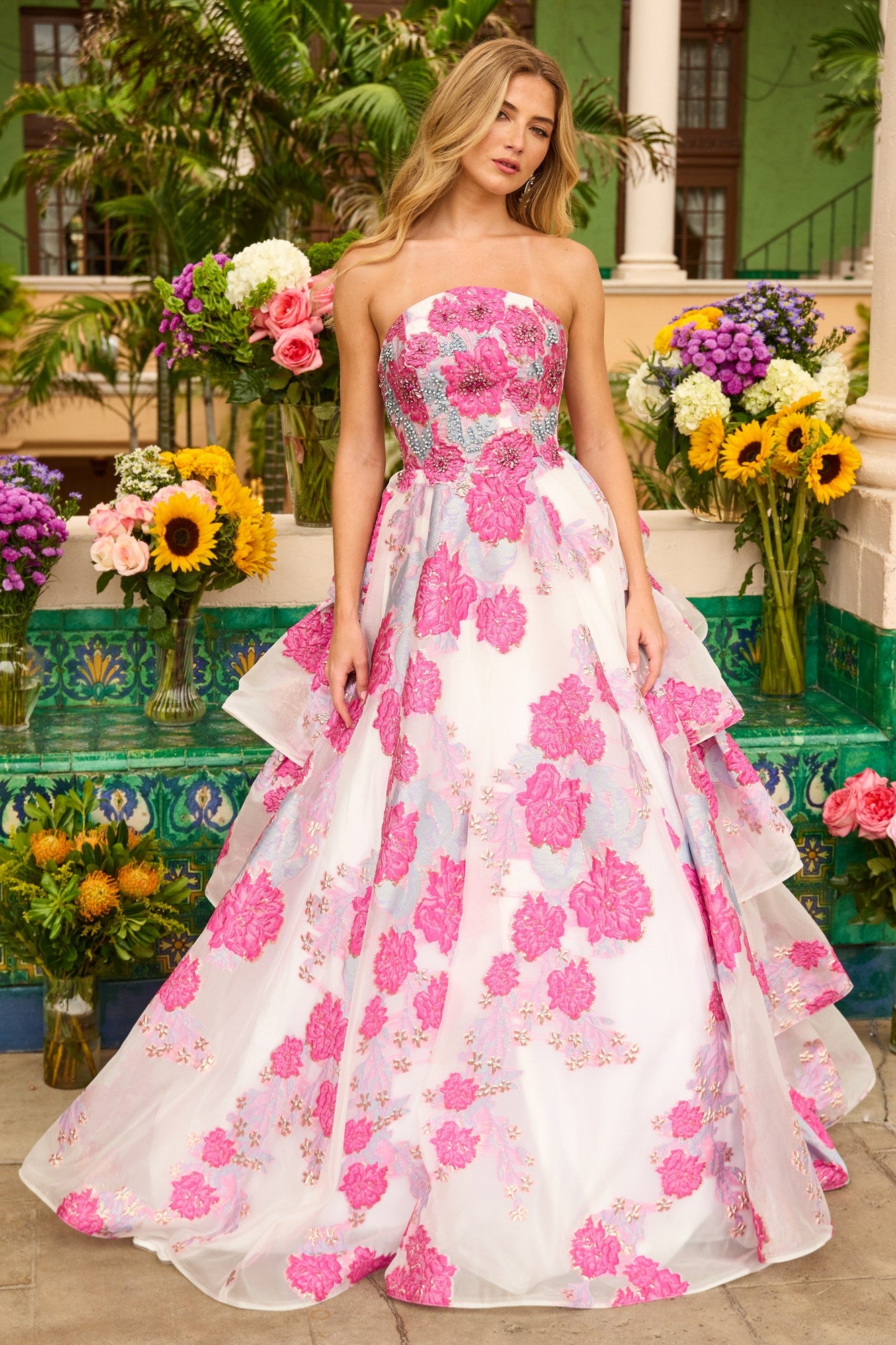A woman poses outdoors in the Ava Presley 42292 long prom dress, featuring a strapless design, bold pink floral patterns, and a tiered ruffle back, surrounded by colorful flowers and lush greenery.